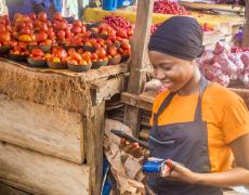 An image showing a woman making a sales transaction on her phone at a vegetable market. 