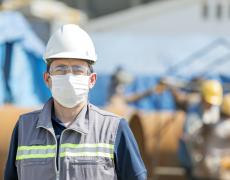 A man wears a hard hat, face mask, and vest in the foreground of a factory